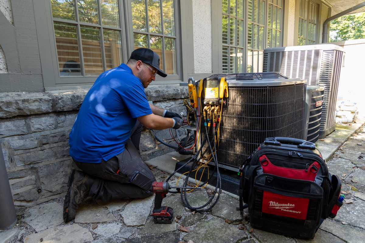 HVAC technician kneeling beside a residential air conditioning unit, using diagnostic tools to perform maintenance, with a tool bag and power drill nearby on a stone patio.