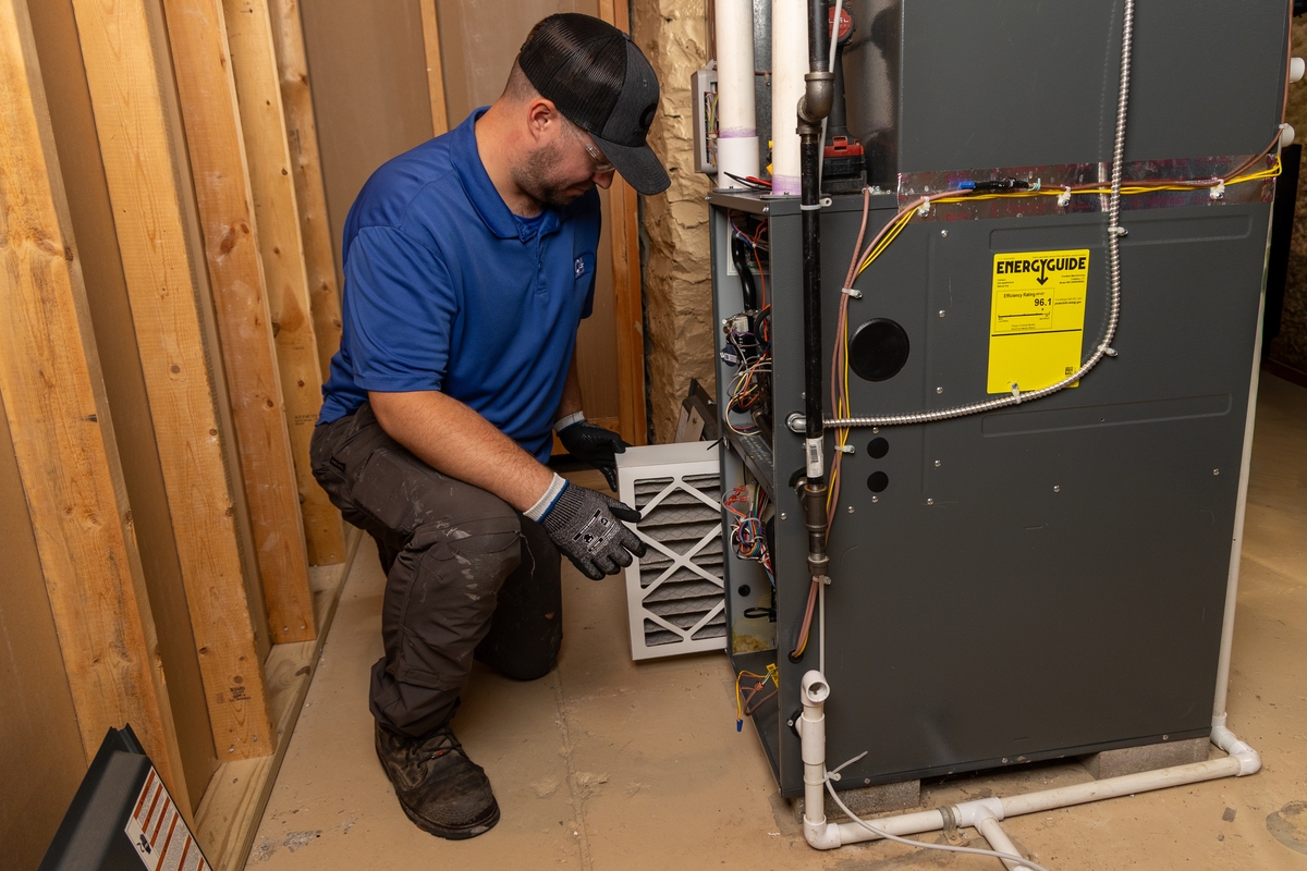 HVAC technician kneeling next to a residential furnace inside a utility room, inspecting or replacing a dirty air filter during maintenance.