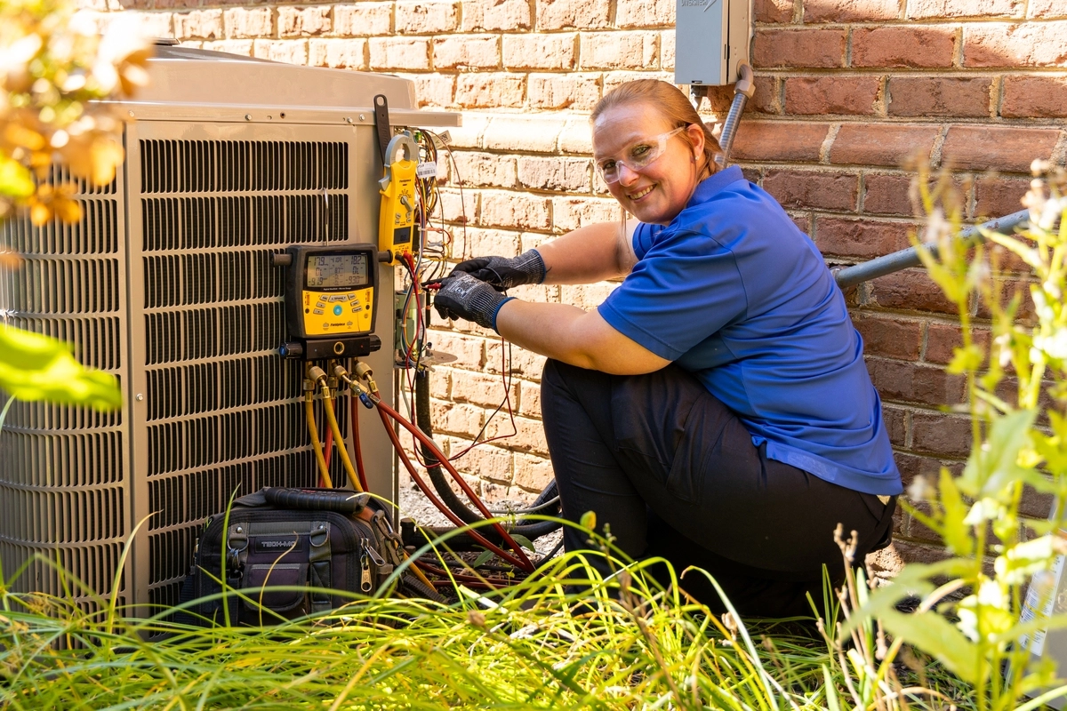 Female HVAC technician smiling while servicing an outdoor air conditioning unit next to a brick home, using digital gauges and tools surrounded by overgrown greenery.