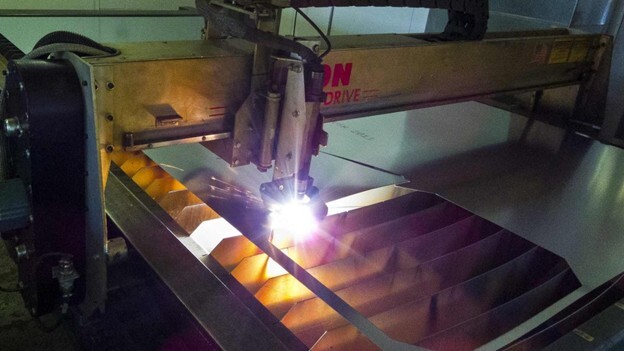 Close-up view of a CNC plasma cutting machine in operation, emitting a bright plasma arc while cutting through a sheet of metal. The machine is mounted on a rail system and cuts with high precision. Metal slats beneath the cutting area catch the molten material.