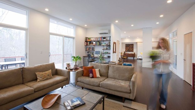 Modern, open-concept living room with neutral-colored furniture, large windows letting in natural light, and recessed lighting in the ceiling. Two beige sofas surround a coffee table with decor and books. Shelving with books and plants is seen in the background. A woman with motion blur is walking through the space holding a potted plant. The room leads into a dining and kitchen area in the background.