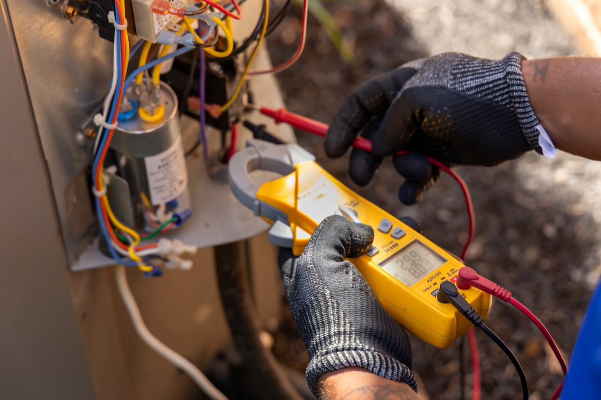 Close-up of an HVAC technician's gloved hands using a digital multimeter to test electrical components and wiring on a residential air conditioning unit.