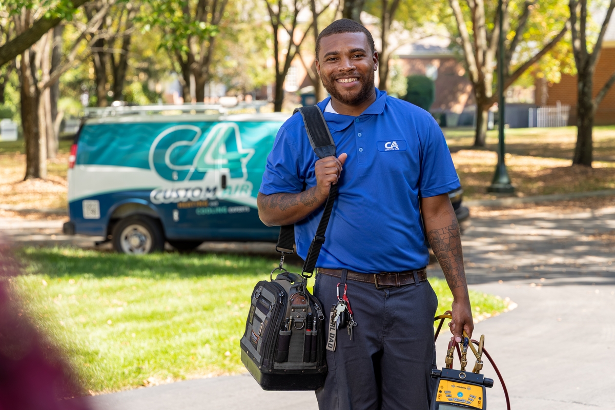 Smiling HVAC technician standing outside with tools in hand and a shoulder bag, wearing a blue Custom Air uniform, with a branded service van parked on a residential street in the background.