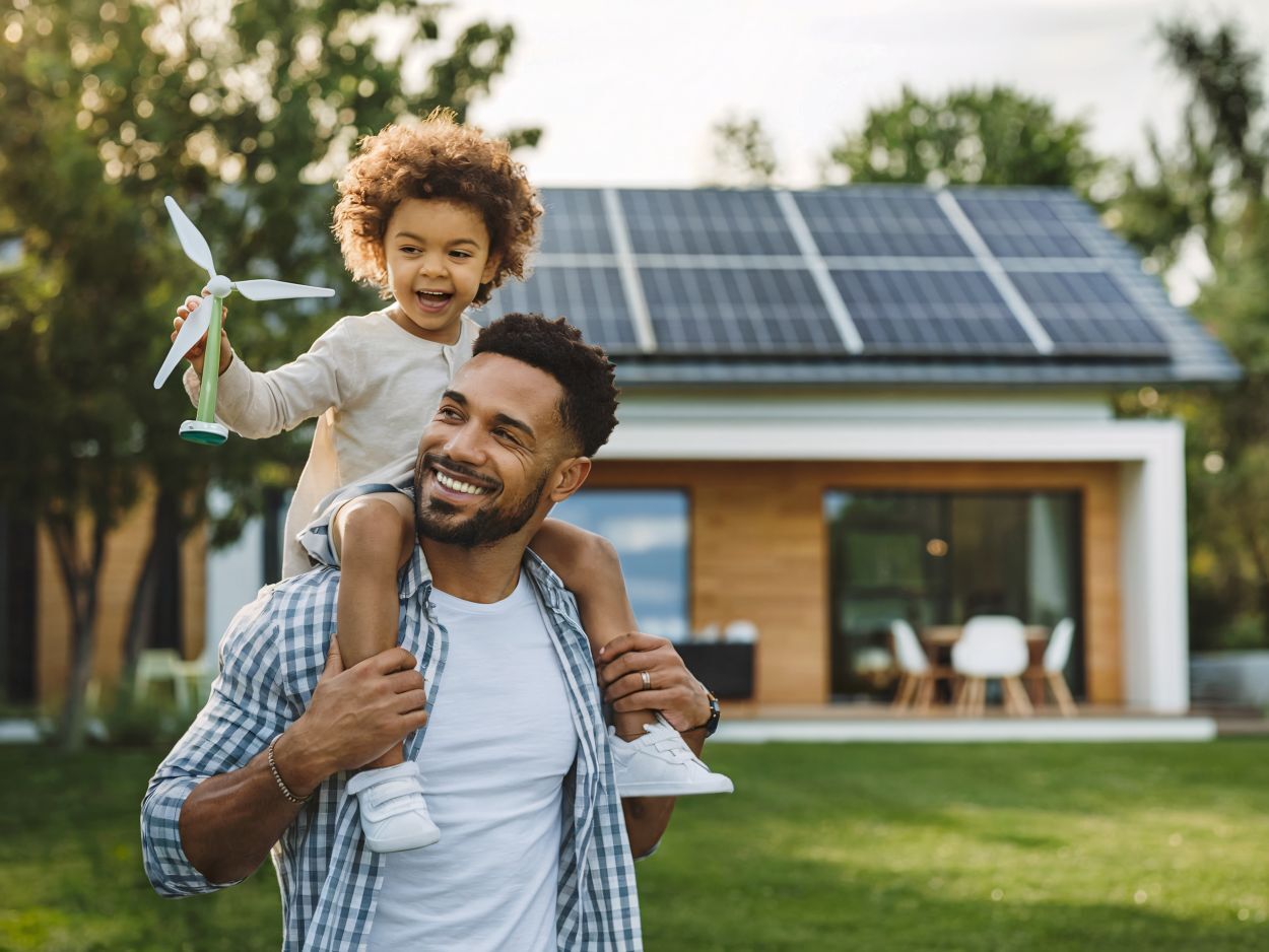 A father carries his child on his shoulders in a yard with a modern house featuring rooftop solar panels
