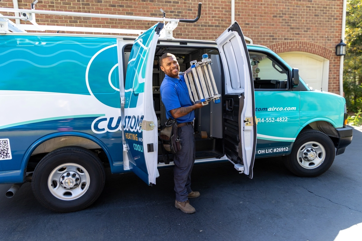 HVAC technician smiling while unloading a foldable ladder from a Custom Air service van parked in a residential driveway, with garage and brick wall in the background.