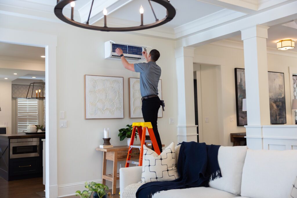 HVAC technician standing on a ladder servicing a wall-mounted ductless mini-split unit in a modern, well-lit living room.