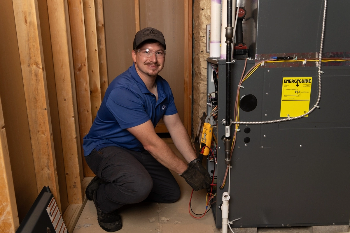 HVAC technician smiling at the camera while performing electrical diagnostics on a residential furnace unit inside a utility room, wearing safety glasses and gloves.