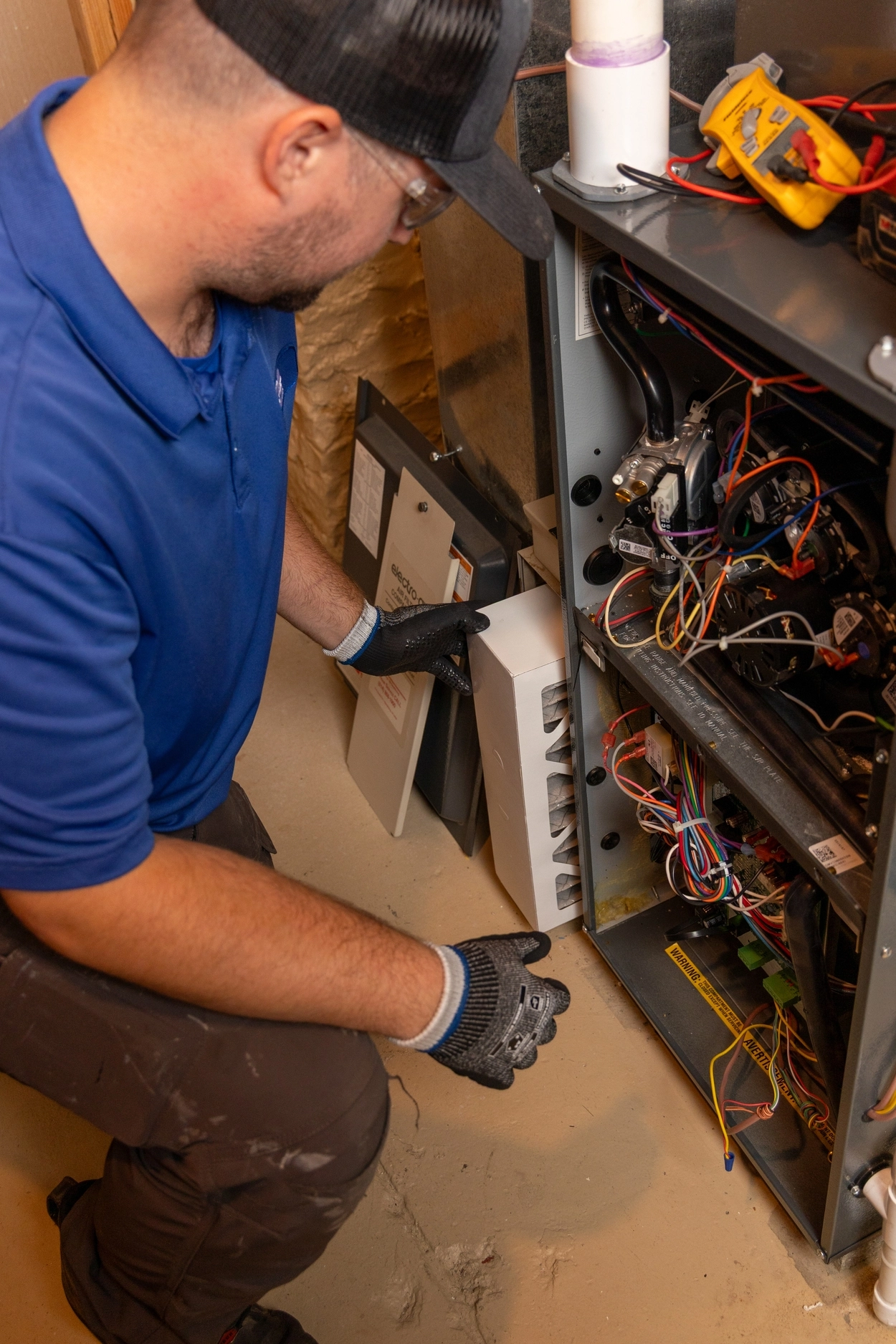 HVAC technician installing or inspecting an air purifier component inside a residential furnace unit during maintenance, with tools and wiring visible around the open panel.