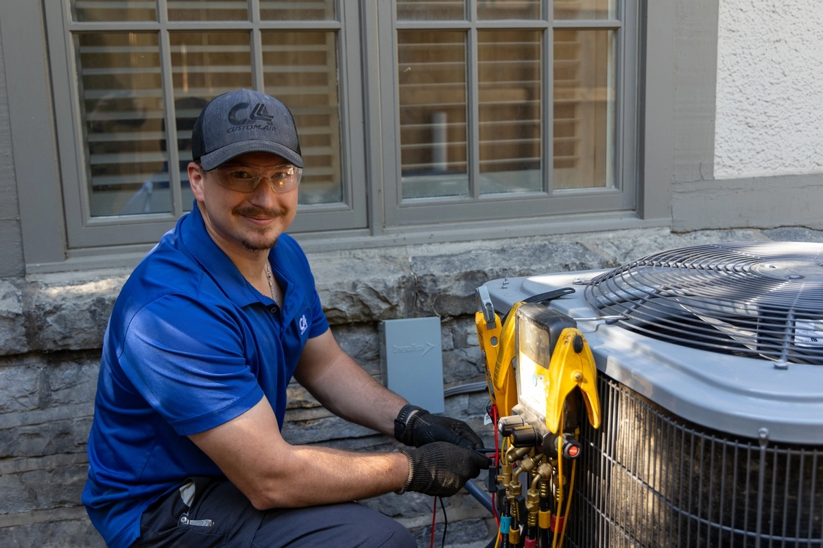HVAC technician in a blue uniform smiling while working on a residential air conditioning unit outside a home, using digital diagnostic tools to check system performance.