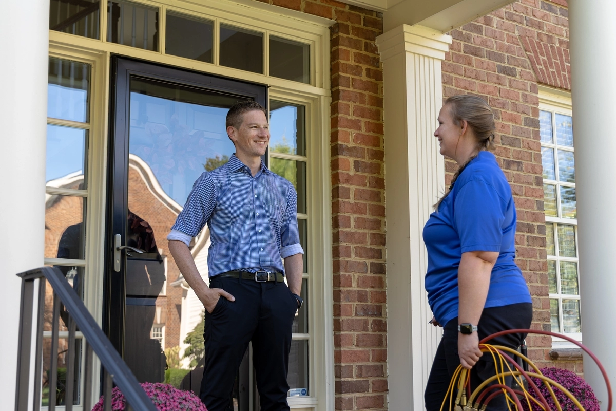 Female HVAC technician speaking with a smiling homeowner at the front door of a brick house, holding diagnostic hoses while standing on the front porch.