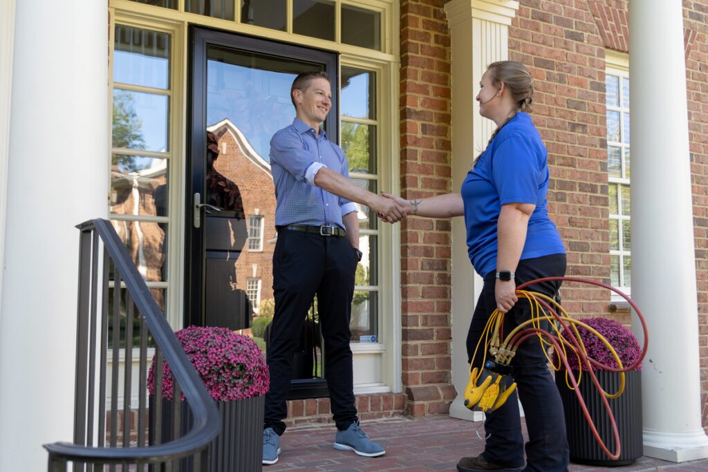 HVAC technician shaking hands with a smiling homeowner on the front porch of a brick house, holding diagnostic gauges in one hand, signifying the completion of a service visit.
