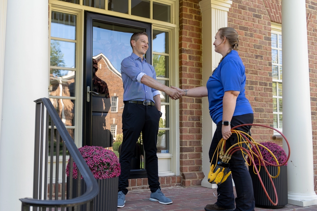 HVAC technician shaking hands with a smiling homeowner on the front porch of a brick house, holding diagnostic gauges in one hand, signifying the completion of a service visit.