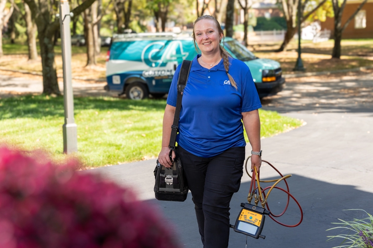 Female HVAC technician walking up a residential driveway with a tool bag and diagnostic gauges in hand, smiling with a branded service van parked in the background.