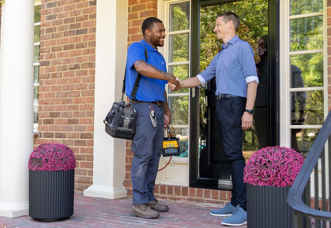HVAC technician shaking hands with a homeowner on the front porch of a brick house after completing a service call, holding diagnostic tools and a tool bag.