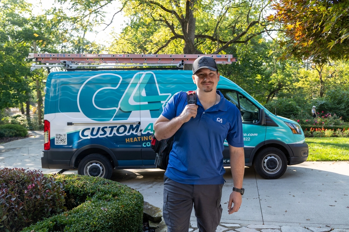 HVAC technician standing in front of a Custom Air service van in a residential driveway, wearing a blue uniform and carrying a tool backpack, ready for a service visit.
