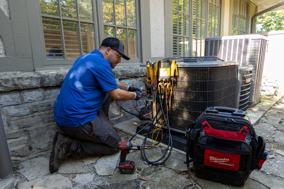 HVAC technician kneeling beside a residential air conditioning unit, using diagnostic tools to perform maintenance, with a tool bag and power drill nearby on a stone patio.