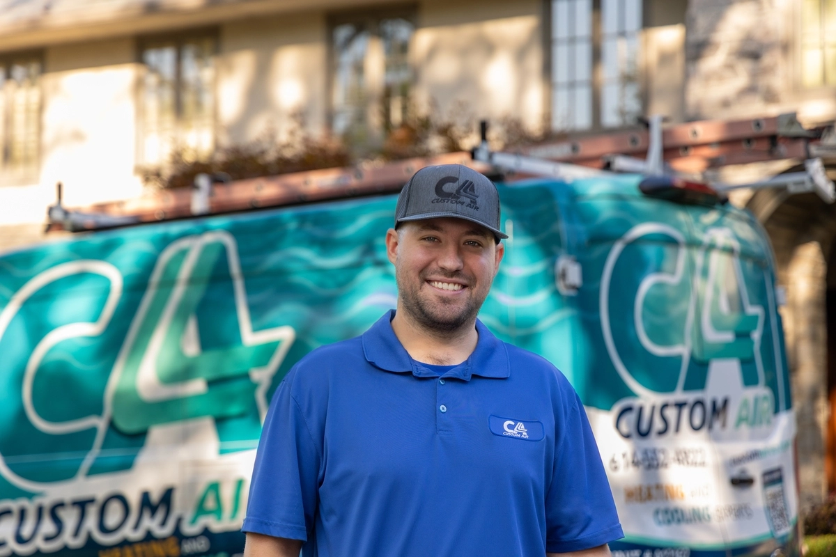 Smiling HVAC technician standing in front of a Custom Air service van, wearing a blue uniform and cap, ready for a residential service call with a home in the background.