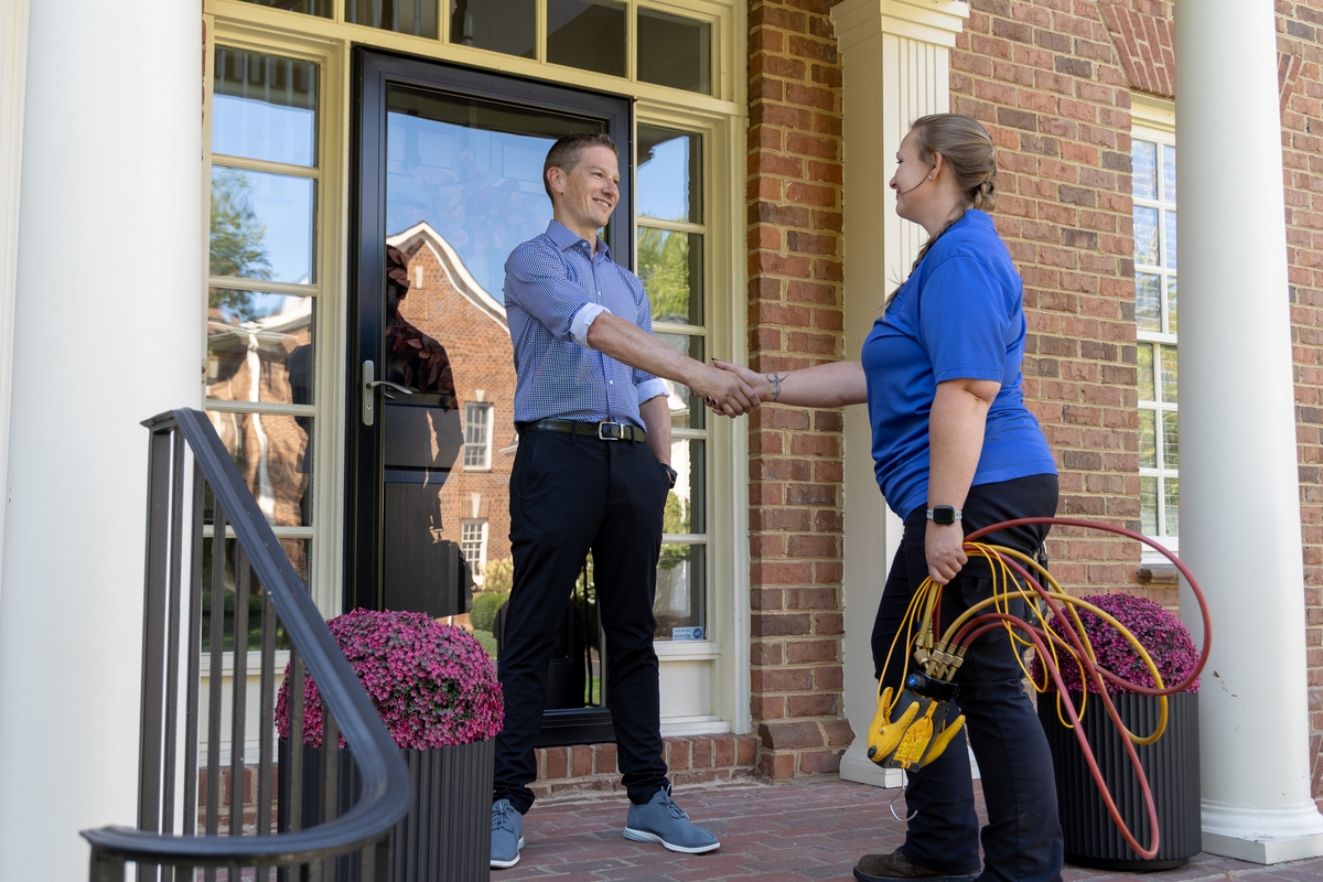 HVAC technician shaking hands with a smiling homeowner on the front porch of a brick house, holding diagnostic gauges in one hand, signifying the completion of a service visit.