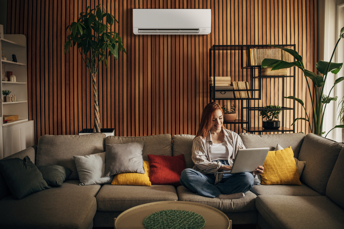 Young woman sitting on a cozy sectional sofa using a laptop in a modern living room with warm wood paneling, colorful pillows, and a ductless mini-split air conditioner mounted on the wall above.