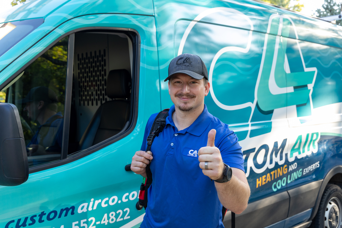 HVAC technician in a blue uniform giving a thumbs-up while standing in front of a Custom Air service van, smiling confidently with a tool backpack over one shoulder.