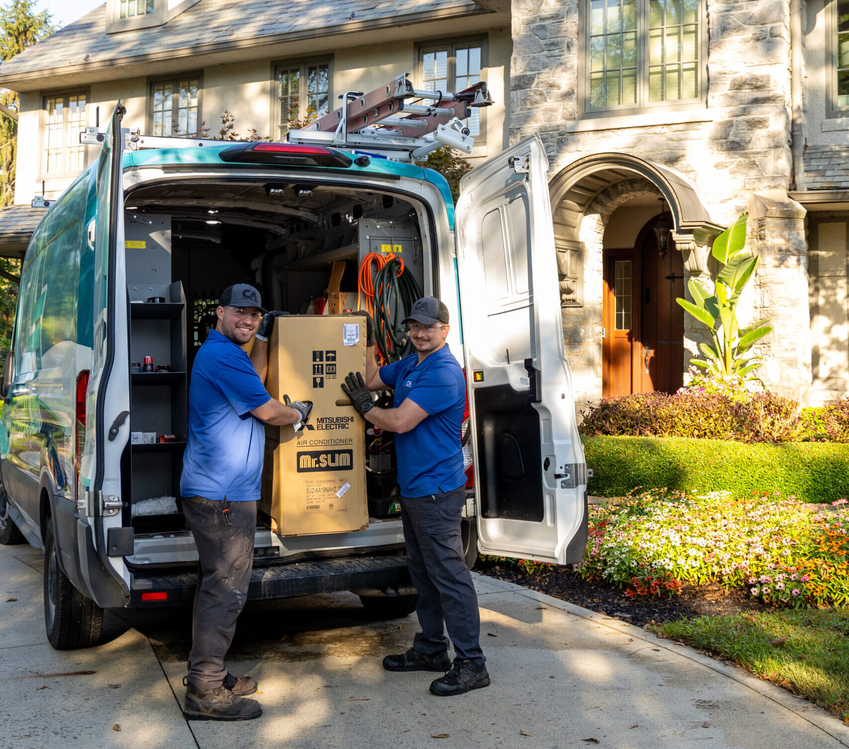 Two HVAC technicians unloading a large unit from the back of a service van in the driveway of a large upscale home, preparing for an installation on a sunny day.