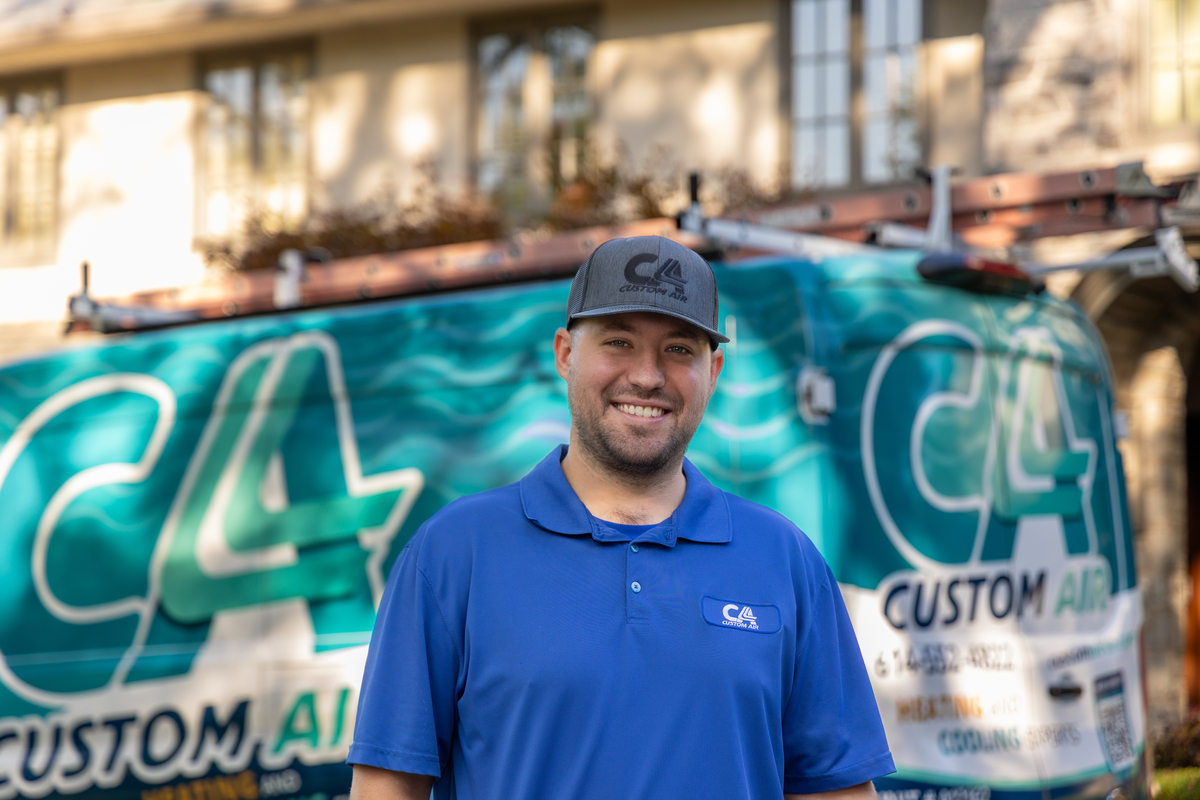 Smiling HVAC technician standing in front of a Custom Air service van, wearing a blue uniform and cap, ready for a residential service call with a home in the background.