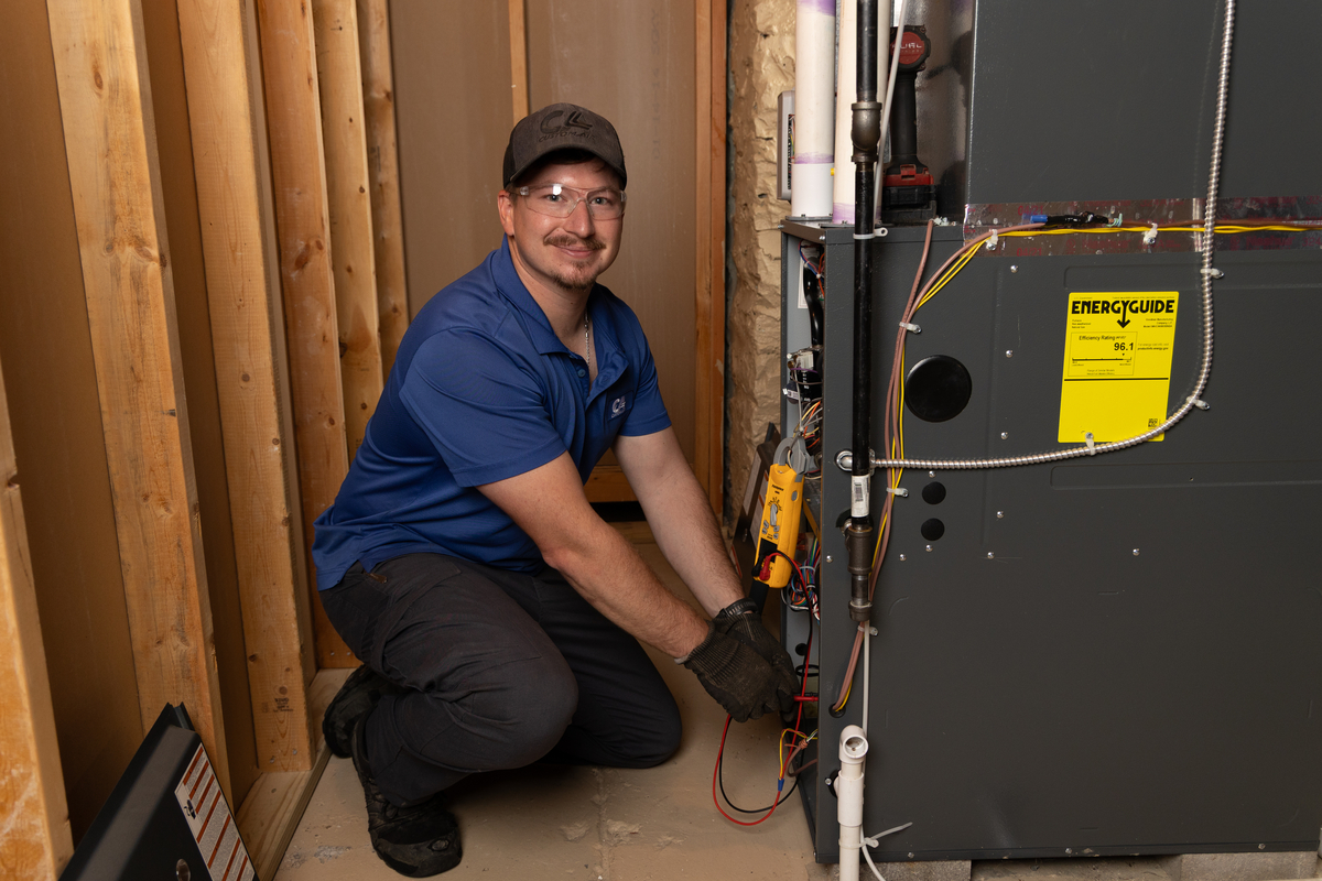 HVAC technician smiling at the camera while performing electrical diagnostics on a residential furnace unit inside a utility room, wearing safety glasses and gloves.