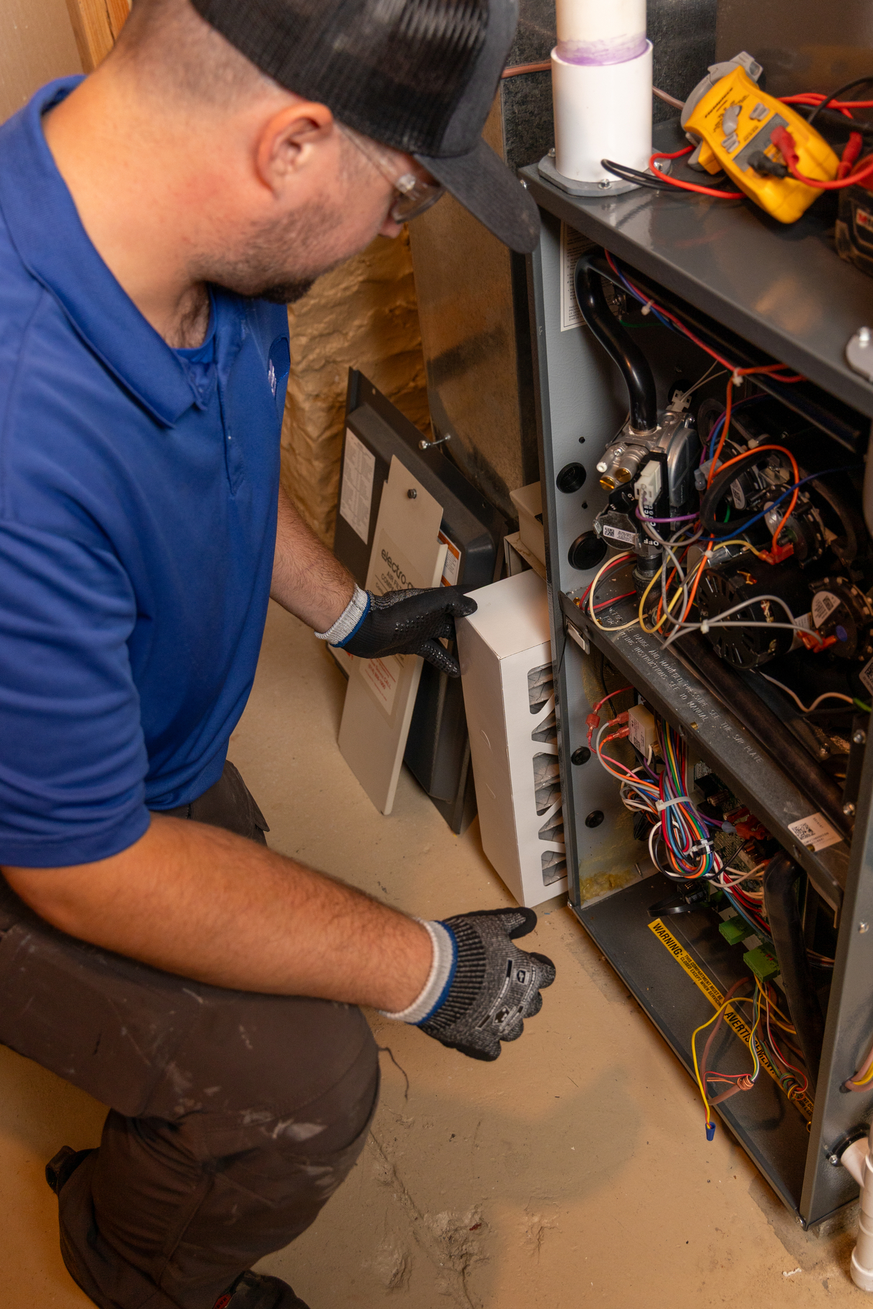 HVAC technician installing or inspecting an air purifier component inside a residential furnace unit during maintenance, with tools and wiring visible around the open panel.