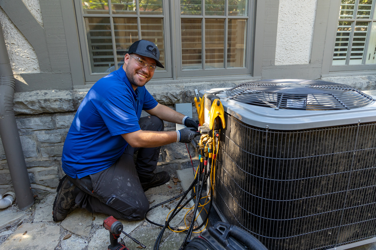 HVAC technician kneeling beside an outdoor residential AC unit, smiling while using digital diagnostic gauges and tools to perform system maintenance.
