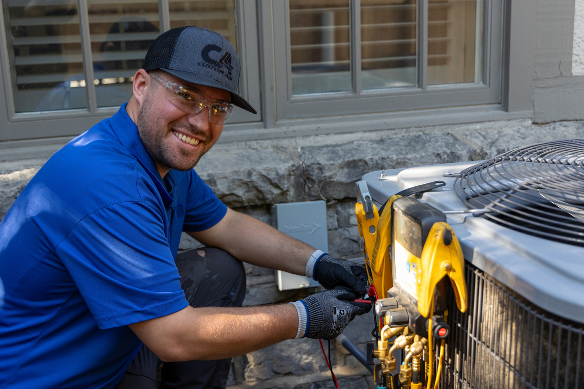 Smiling HVAC technician wearing safety glasses and gloves while performing diagnostics on a residential air conditioning unit with digital gauges outside a home.