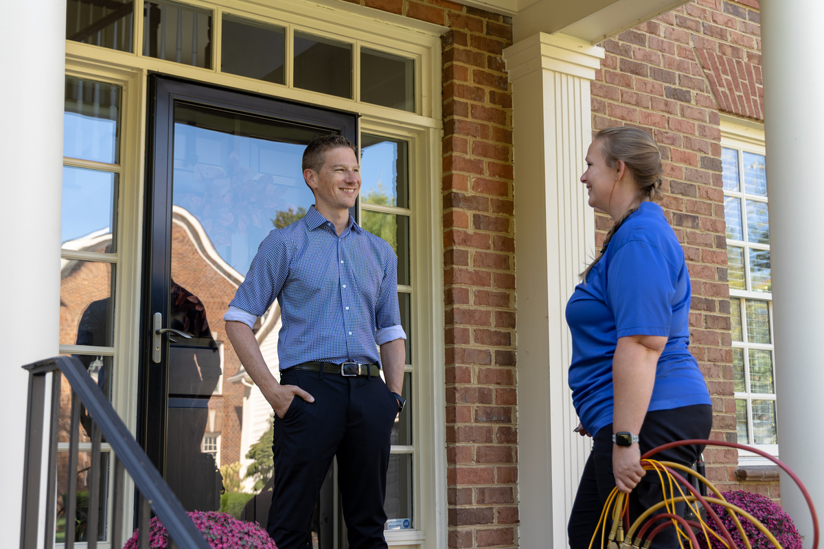 Female HVAC technician speaking with a smiling homeowner at the front door of a brick house, holding diagnostic hoses while standing on the front porch.
