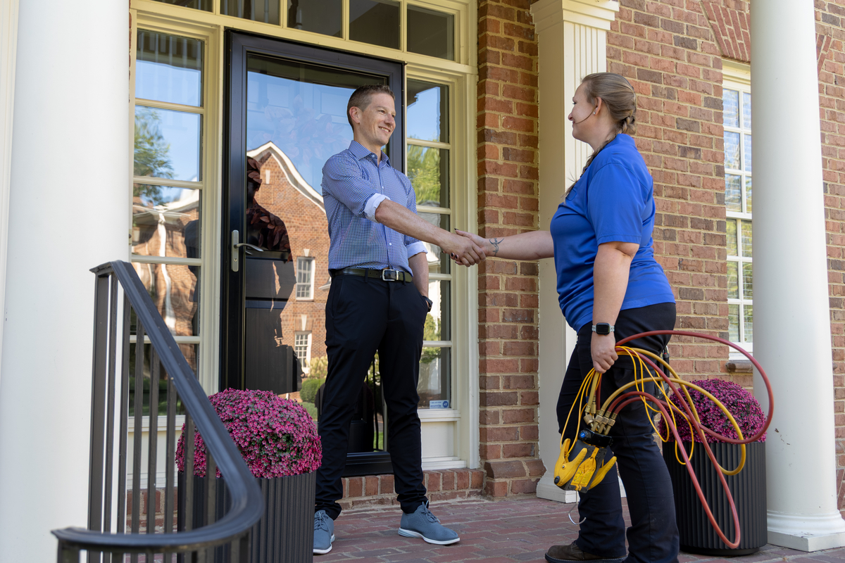 HVAC technician shaking hands with a smiling homeowner on the front porch of a brick house, holding diagnostic gauges in one hand, signifying the completion of a service visit.