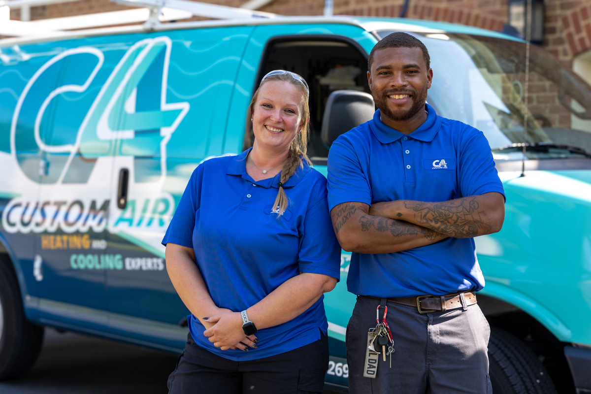Two HVAC technicians, a woman and a man, standing confidently and smiling in front of a Custom Air service van, both wearing blue uniforms, ready for their next job.