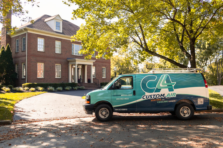 Custom Air HVAC service van parked in the driveway of a large brick residential home on a sunny day, surrounded by mature trees and landscaped greenery.